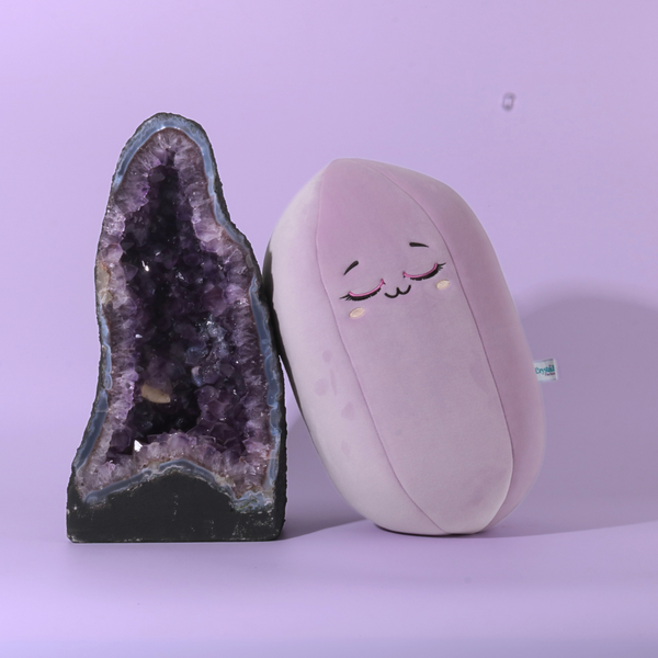 Peluche cristal rose en forme de cristal avec visage souriant à côté d'une géode d'améthyste sur fond violet clair