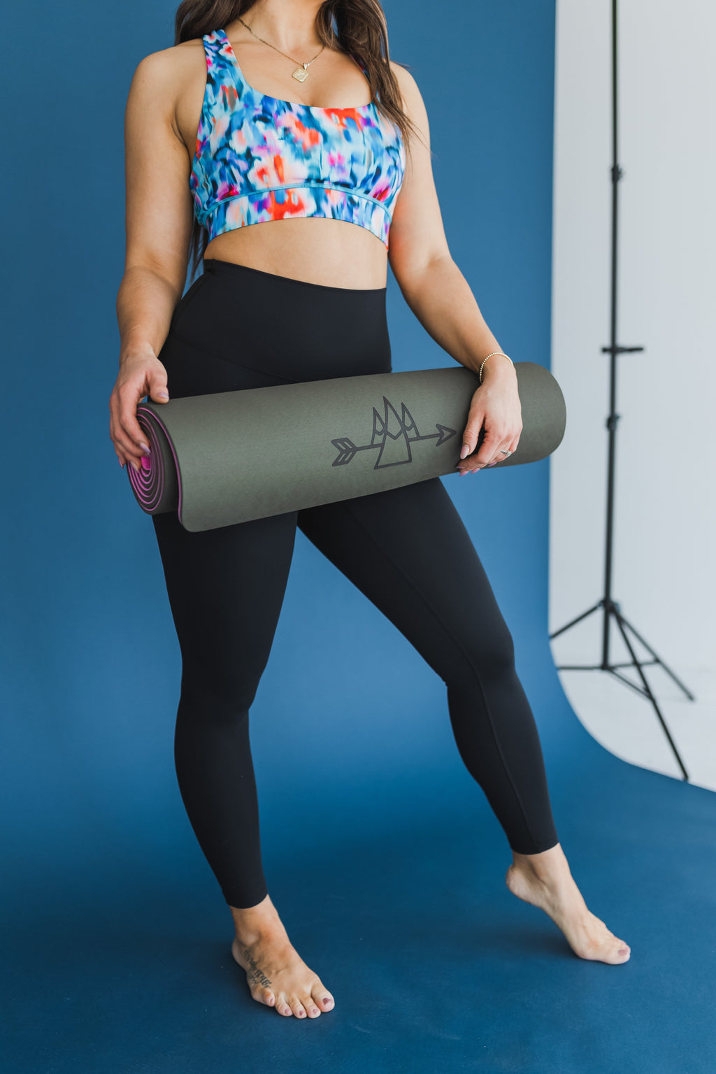 Femme tenant un tapis de yoga enroulé vert foncé devant un fond bleu